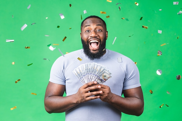 Gelukkige winnaar! Jonge rijke Afro-Amerikaanse zwarte man in casual t-shirt met geld dollarbiljetten met verrassing geïsoleerd over groene achtergrond.