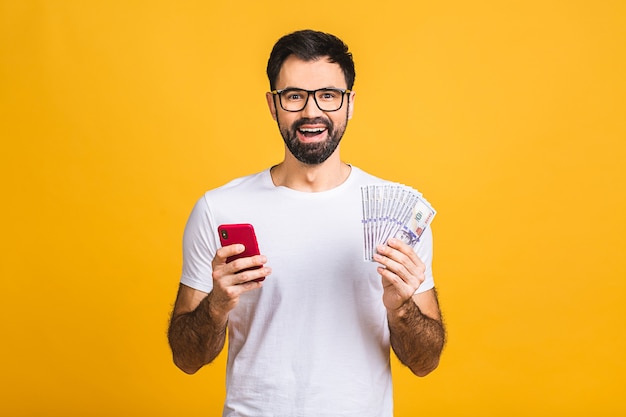 Gelukkige winnaar! opgewonden jonge bebaarde man in casual met veel geld in dollar valuta en mobiele telefoon in handen geïsoleerd over gele muur.