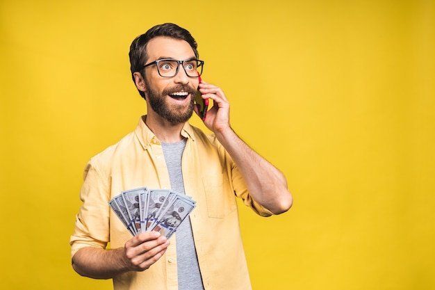 Gelukkige winnaar! Opgewonden jonge, bebaarde man in casual met veel geld in dollarvaluta's en het gebruik van mobiele telefoon in handen geïsoleerd op gele achtergrond.