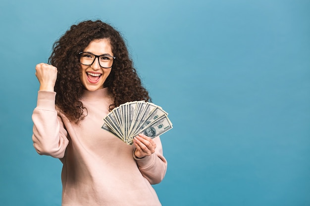 Gelukkige winnaar! Portret van een vrolijke jonge krullende vrouw met geld bankbiljetten en vieren geïsoleerd op blauwe achtergrond.