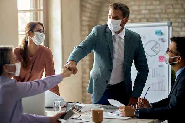 Gelukkige zakelijke collega's met gezichtsmaskers die tegen elkaar stoten na een succesvolle bijeenkomst op kantoor