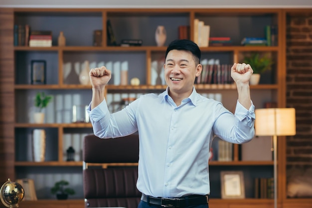 Gelukkige zakenman die danst en de overwinning viert, zittend aan een tafel in een klassiek kantoor-Aziatische werken op de computer