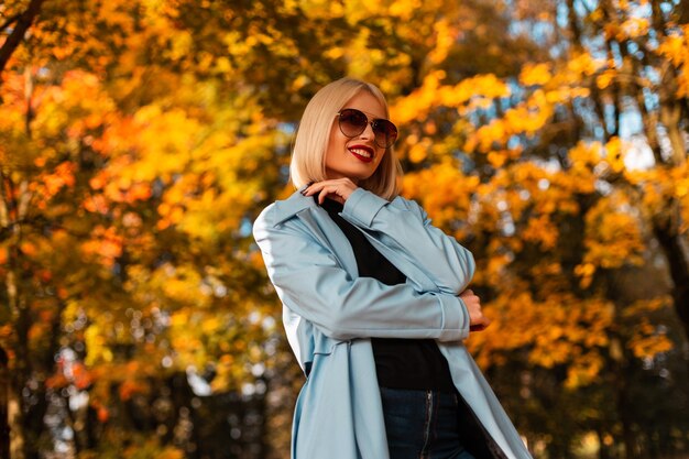 Gelukkige zakenvrouw met een glimlach in modieuze kleding met zonnebril loopt in het park met felgeel herfstgebladerte