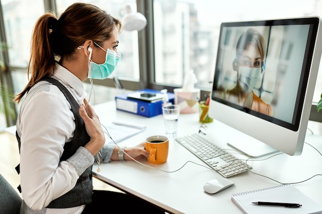 Gelukkige zakenvrouw met gezichtsmasker die via de computer met haar collega communiceert terwijl ze een koffiepauze op kantoor heeft