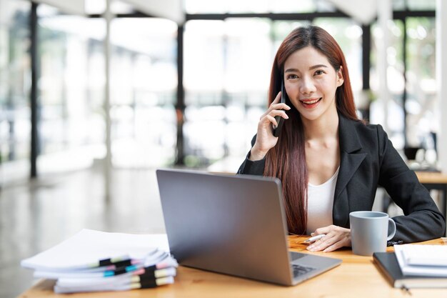 Gelukkige zakenvrouw zit aan een bureau achter haar laptop en praat met iemand op haar mobiele telefoon terwijl ze op kantoor werkt