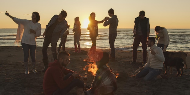 Gelukkige zorgeloze jonge vrienden die plezier hebben en bier drinken bij een kampvuur op het strand terwijl de zon ondergaat