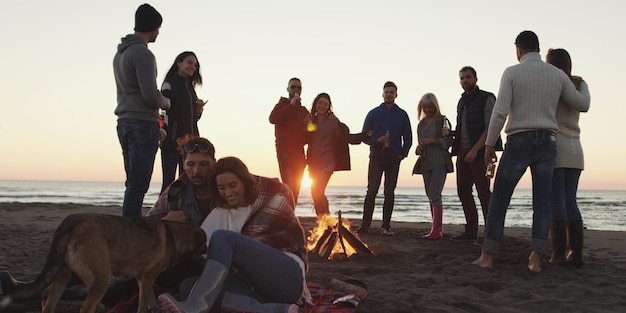 Gelukkige zorgeloze jonge vrienden die plezier hebben en bier drinken bij een kampvuur op het strand terwijl de zon ondergaat