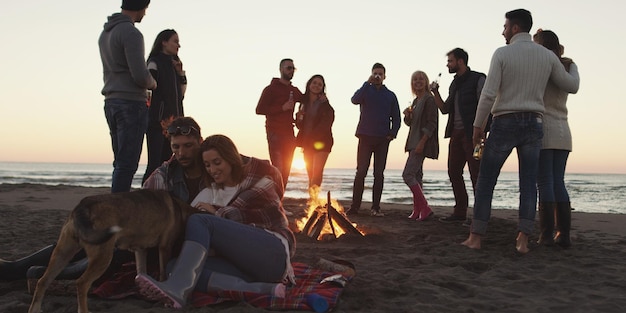 Gelukkige zorgeloze jonge vrienden die plezier hebben en bier drinken bij een kampvuur op het strand terwijl de zon ondergaat