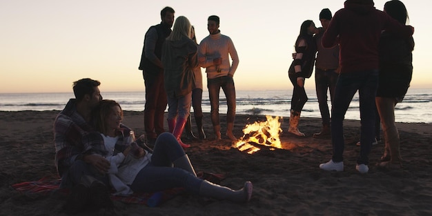 Gelukkige zorgeloze jonge vrienden die plezier hebben en bier drinken bij een kampvuur op het strand terwijl de zon ondergaat