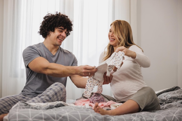 Gelukkige zwangere paar kijken naar babykleertjes terwijl u ontspant op een bed in de slaapkamer.