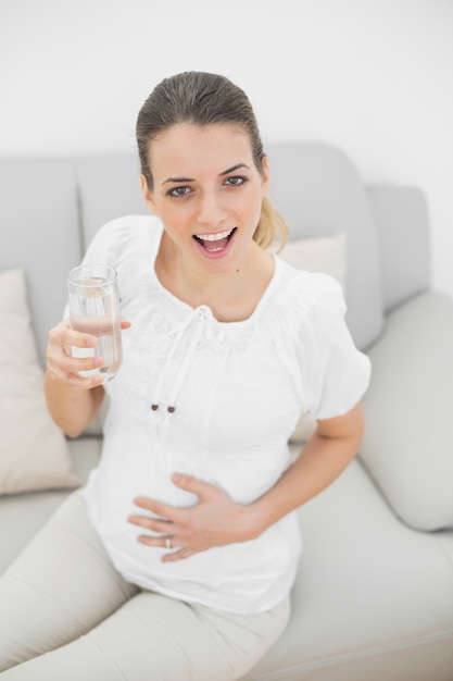 Gelukkige zwangere vrouw die een glas water houdt dat bij camera glimlacht