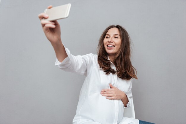 Gelukkige zwangere vrouw maakt selfie in studio geïsoleerde grijze achtergrond