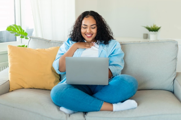 Gelukkige zwarte extra grote dame die thuis een laptop gebruikt om nieuws te lezen of een videogesprek te voeren met vrienden die thuis op de bank rusten