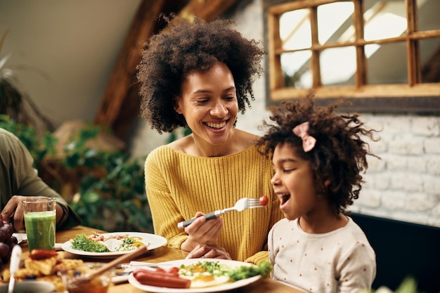 Gelukkige zwarte moeder die haar dochter voedt tijdens een maaltijd aan de eettafel