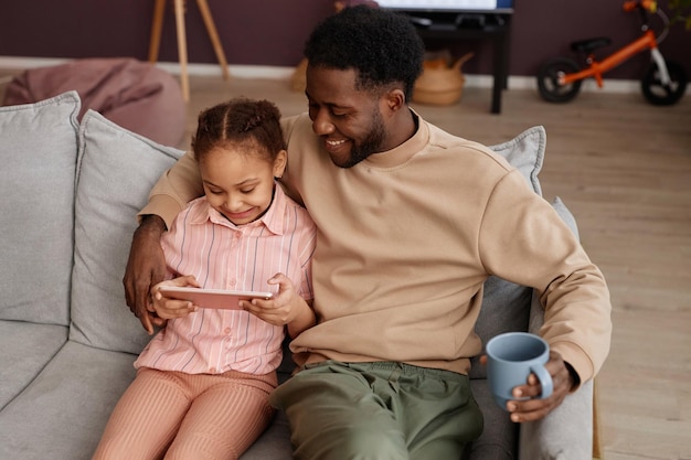 Gelukkige zwarte vader met klein meisje met behulp van tablet zitten op de bank thuis en