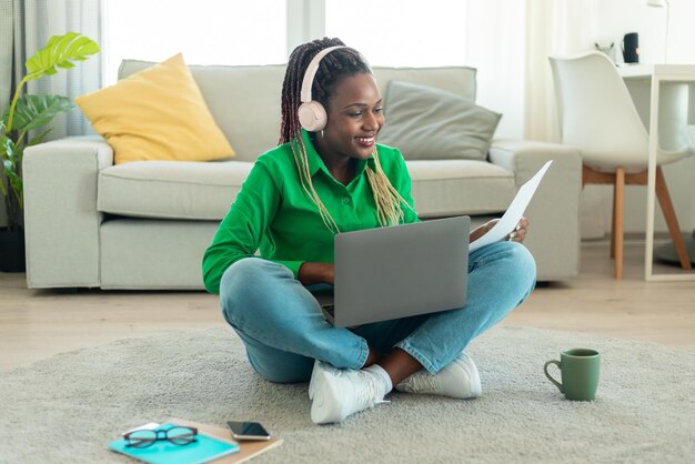 Gelukkige zwarte vrouw die een laptop gebruikt en papieren leest die online werkt of online studeert vanuit huis