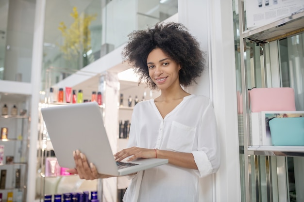 Geluksdag. Glimlachende jonge vrouw met donker krullend haar die zich met laptop in zaal met kosmetische producten bevinden