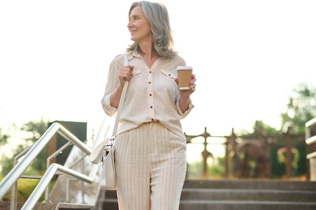 Geluksdag. Vrij volwassen elegante vrouw met koffie op zoek naar kant op trappen in park op zomerdag