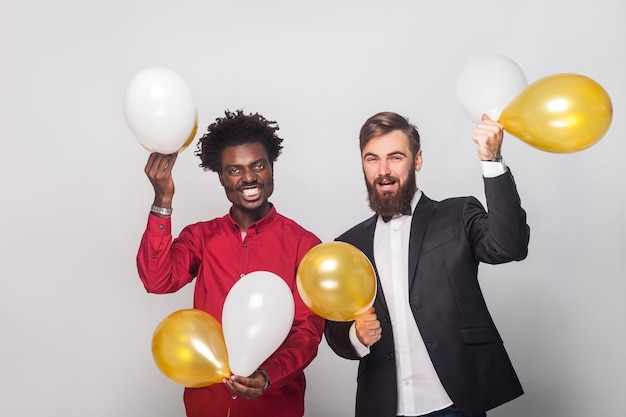 Geluksvrienden die verjaardag vieren, gouden en witte luchtballon vasthouden, handen omhoog en glimlachen. Studio opname, grijze muur