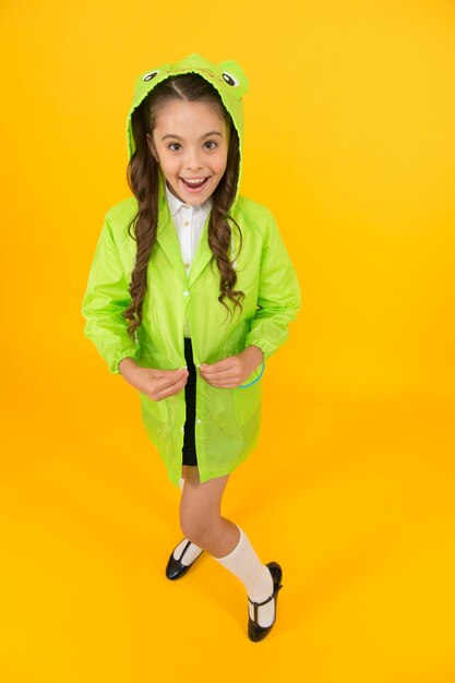 Gemakkelijk vast te maken gelukkig schoolmeisje gaat uit op gele achtergrond klein schoolmeisje kleedt regenjas en capuchon aan voor regenachtige dag klein schoolmeisje voelt zich beschermd voor herfstweer