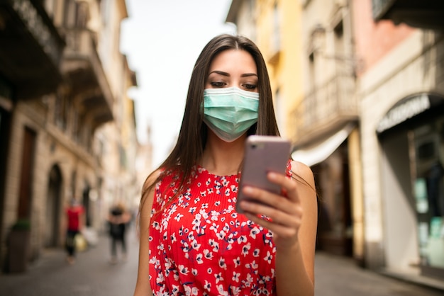 Gemaskerde jonge vrouw die in een stad loopt terwijl het gebruiken van haar mobiele telefoon