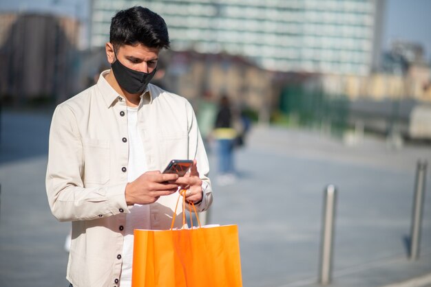 Gemaskerde man met boodschappentassen tijdens het gebruik van een smartphone-, covid- en coronavirusconcept