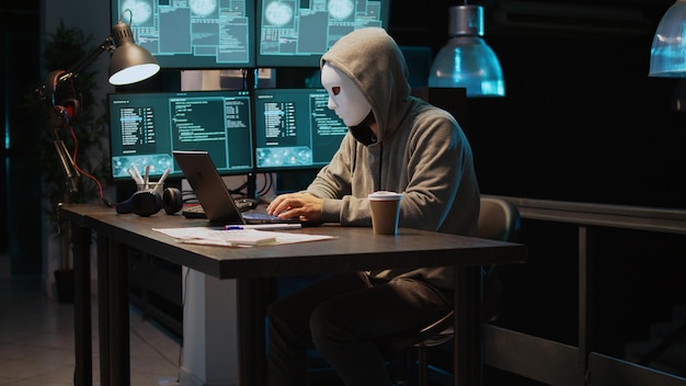 Gemaskerde persoon hackt 's nachts computernetwerk, werkt op laptop en meerdere monitoren om IT-informatie te stelen. Hacker met verborgen identiteit die gevaarlijk handelt, inbreuk op de serverbeveiliging. Schot uit de hand.