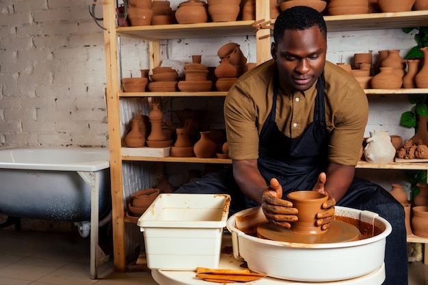 Gemengd ras afro mannelijke pottenbakker met zwarte schort en stijlvol donker shirt zittend aan workshop tafel pottenbakkerswiel glazuur toe te passen op handgemaakte klei bruine pot vaas