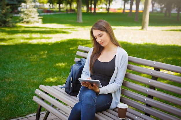 Gemotiveerde Aziatische freelancer in het park. Jonge brunette meisje op een bankje, koffie drinken en online werken met tablet. Lifestyle, technologie en werken op afstand concept