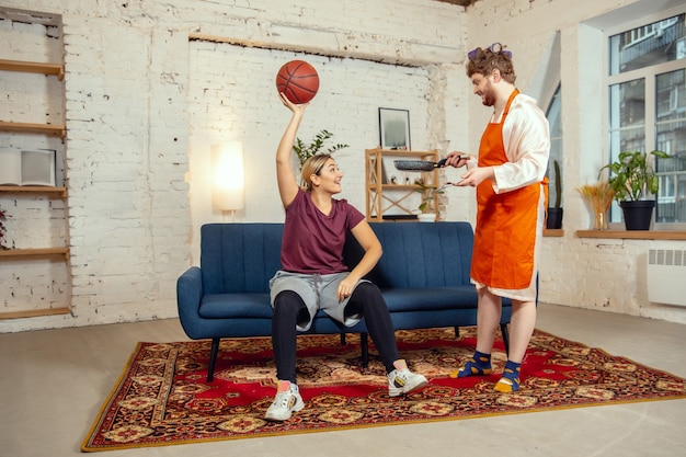 ジェンダーのステレオタイプ。妻と夫は、社会的意味、感覚において、性別としては珍しいことをしています。居間でボールを持ってバスケットボールでトレーニングしている女性が夕食を作っている男性。
