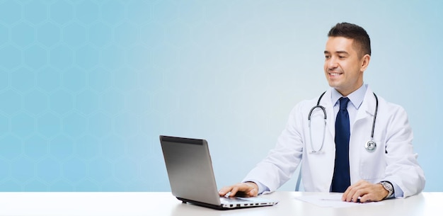 geneeskunde, beroep, technologie en mensen concept - glimlachende mannelijke arts zittend aan tafel met laptop en stethoscoop over blauwe achtergrond