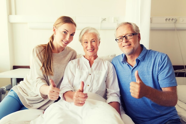 geneeskunde, gezinsondersteuning, gebaar, gezondheidszorg en mensenconcept - gelukkige senior man en jonge vrouw die haar grootmoeder bezoeken en duimen laten zien op de ziekenhuisafdeling