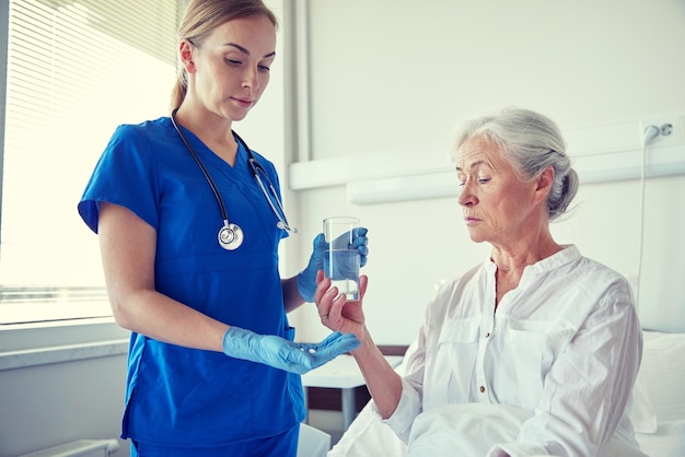 geneeskunde, leeftijd, gezondheidszorg en mensenconcept - verpleegster die medicatie en glas water geeft aan senior vrouw op ziekenhuisafdeling
