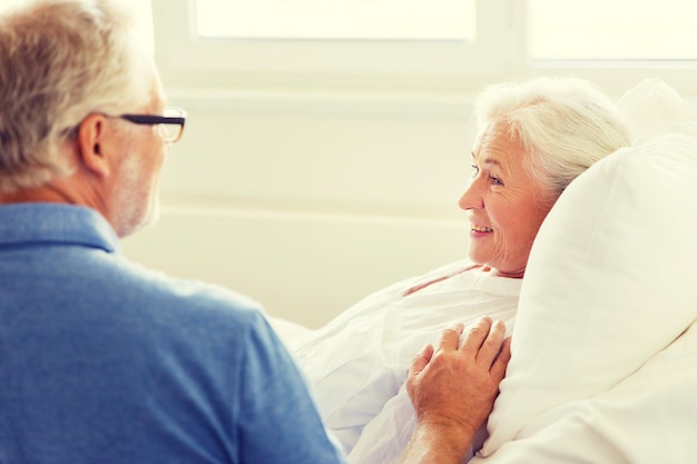 geneeskunde, leeftijd, ondersteuning, gezondheidszorg en mensenconcept - gelukkige senior man die zijn vrouw bezoekt en juicht die in bed ligt op de ziekenhuisafdeling