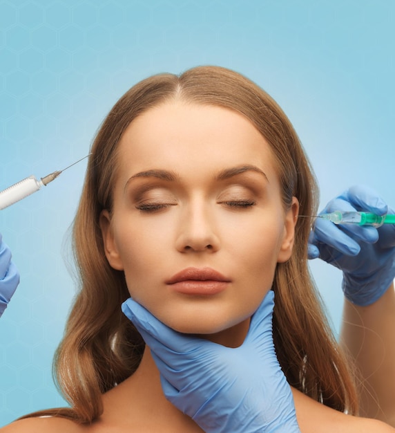 geneeskunde, plastische chirurgie, schoonheid, gezondheid en mensenconcept - handen in medische handschoen met spuit die injectie maken aan mooi vrouwengezicht over blauwe achtergrond