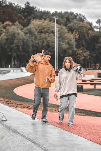 General interest. Girl with wavy hair and red-haired guy with drinks and skateboards talking walking in skate park in afternoon after workout