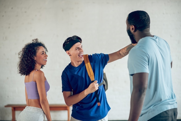 General interest. Two young guys and girl with dark curly hair in sportswear standing communicating indoors