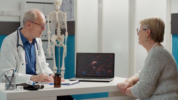 General practitioner explaining coronavirus illustration on\
laptop computer, talking to retired woman in cabinet. covid 19\
virus animation on display at examination checkup appointment.