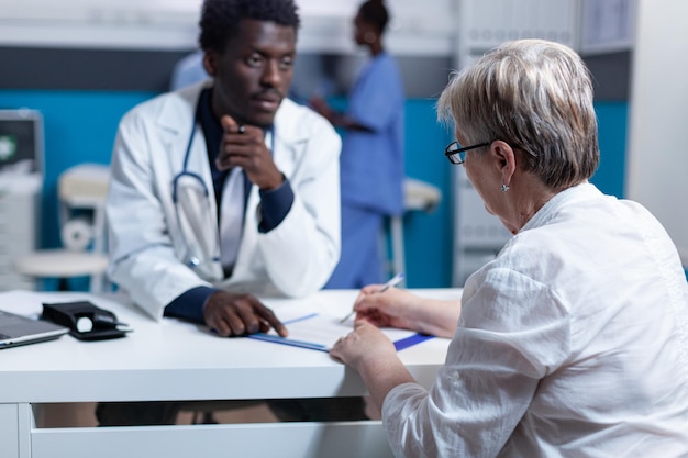 General practitioner explaining effects and side effects of prescribed painkillers to ill senior patient. Elderly woman signing discharge summaries and treatment clinic schedule