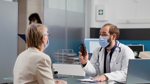 General practitioner giving prescription medicine in bottle to elder patient with disease, attending checkup appointment during pandemic. Senior woman receiving treatment from medic.