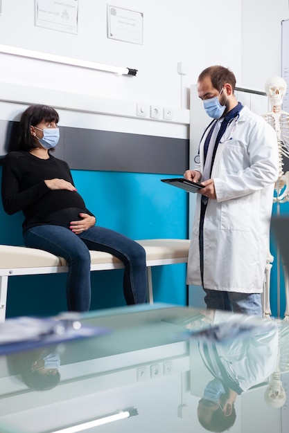 General practitioner holding digital device to consult woman with pregnancy belly, wearing face mask. Specialist using device to do medical consultation with pregnant adult during pandemic.