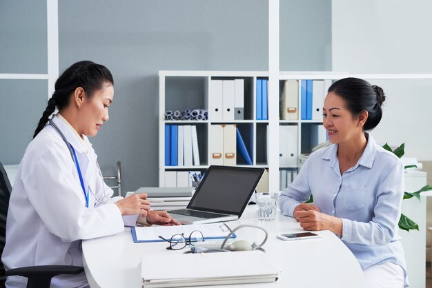 General practitioner talking to senior patient and filling form with her personal data and complaints