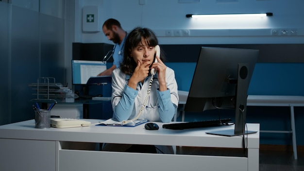 General practitioner woman sitting at desk while talking with\
remote patient using landline phone, discussing medication\
treatment. medical staff working late at night in hospital\
office