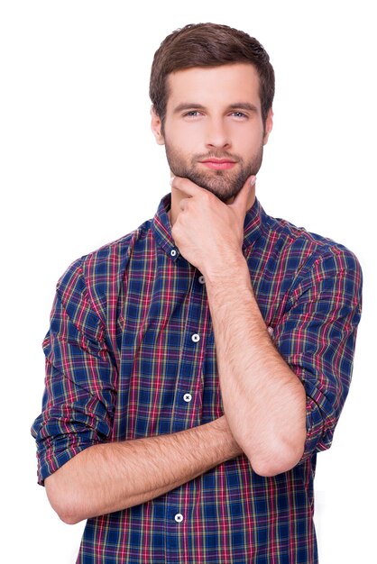 Generating new ideas for you. Thoughtful young man in casual shirt holding hand on chin and looking at camera while standing isolated on white