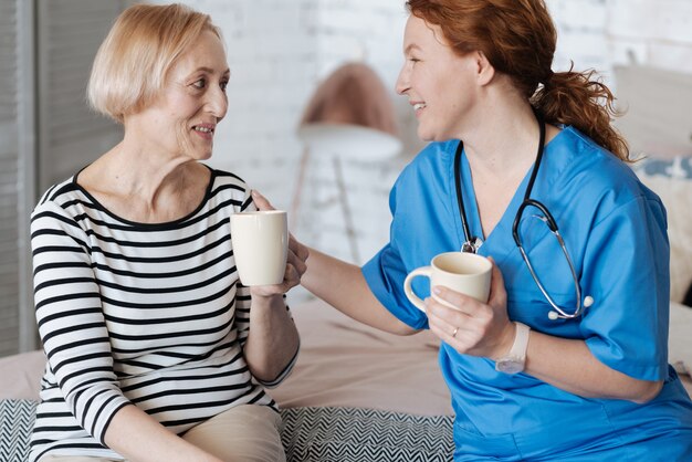 Genezen met positieve emoties. Vriendelijke dankbare rijpe vrouw die de dokter uitnodigt voor een kopje thee nadat ze haar heeft geholpen met de behandeling