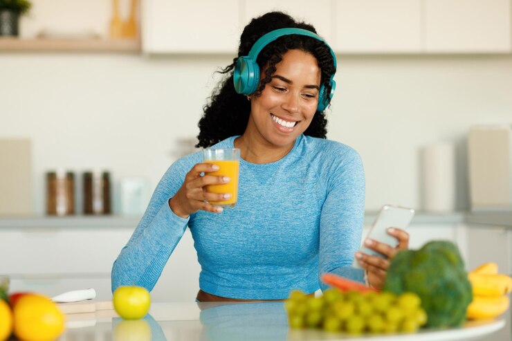 Foto genieten van een gezond drankje gelukkige zwarte dame in sportkleding die zelfgemaakte sap vasthoudt en smartphone gebruikt die in de keuken zit
