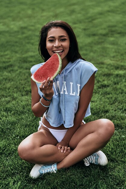 Genieten van een gezonde snack. Aantrekkelijke jonge vrouw die een plakje watermeloen eet en glimlacht terwijl ze op het gras zit