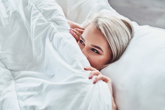 Genieten van een mooie ochtend. Aantrekkelijke jonge vrouw die de helft van haar gezicht bedekt met een deken en glimlacht terwijl ze thuis in bed ligt