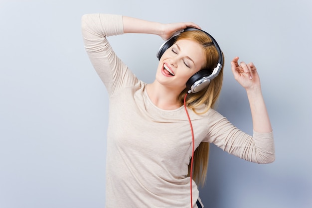 Genieten van haar favoriete liedje. Mooie jonge vrouwen in koptelefoon die ogen gesloten houden en dansen terwijl ze tegen een grijze achtergrond staan