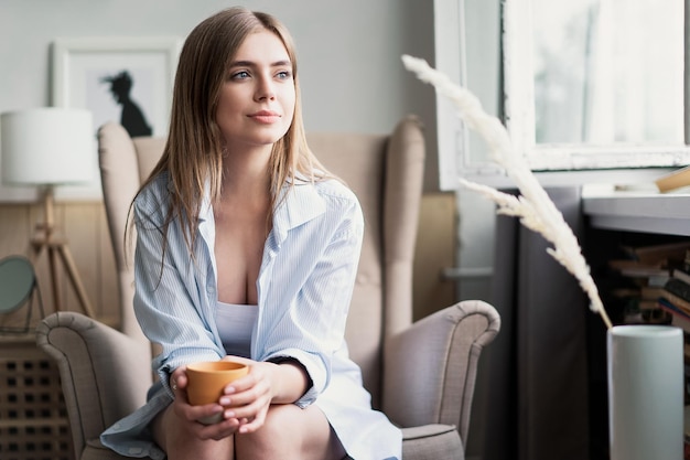 Genieten van haar verse koffie Mooie jonge vrouw die koffie drinkt en door het raam kijkt terwijl ze thuis in een fauteuil zit